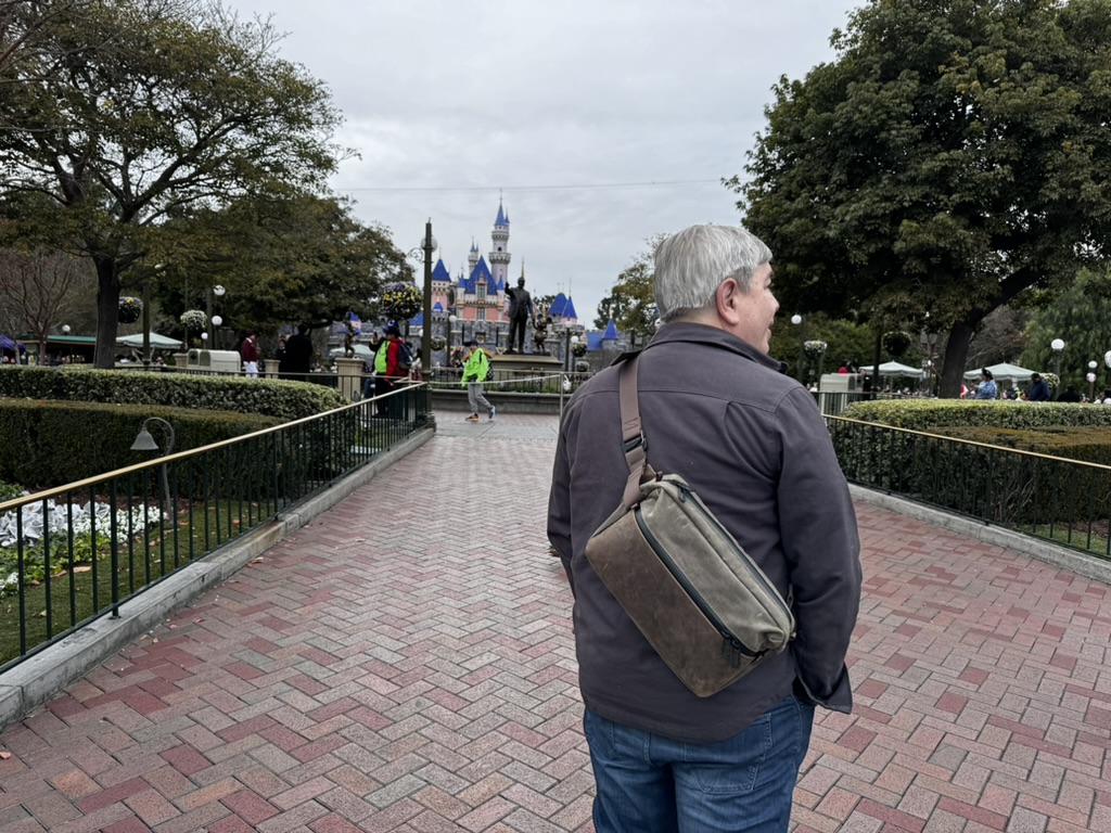 A view from the back of Sparky, walking towards the castle at Disneyland in Anaheim. He's turning his head to the right, is wearing jeans and a brown jacket, and has the Waterfield Hip Sling slung around his left shoulder. It is an overcast day, with green tress showing in front of him, on both sides of the wide pedestrian path he's walking on. 
